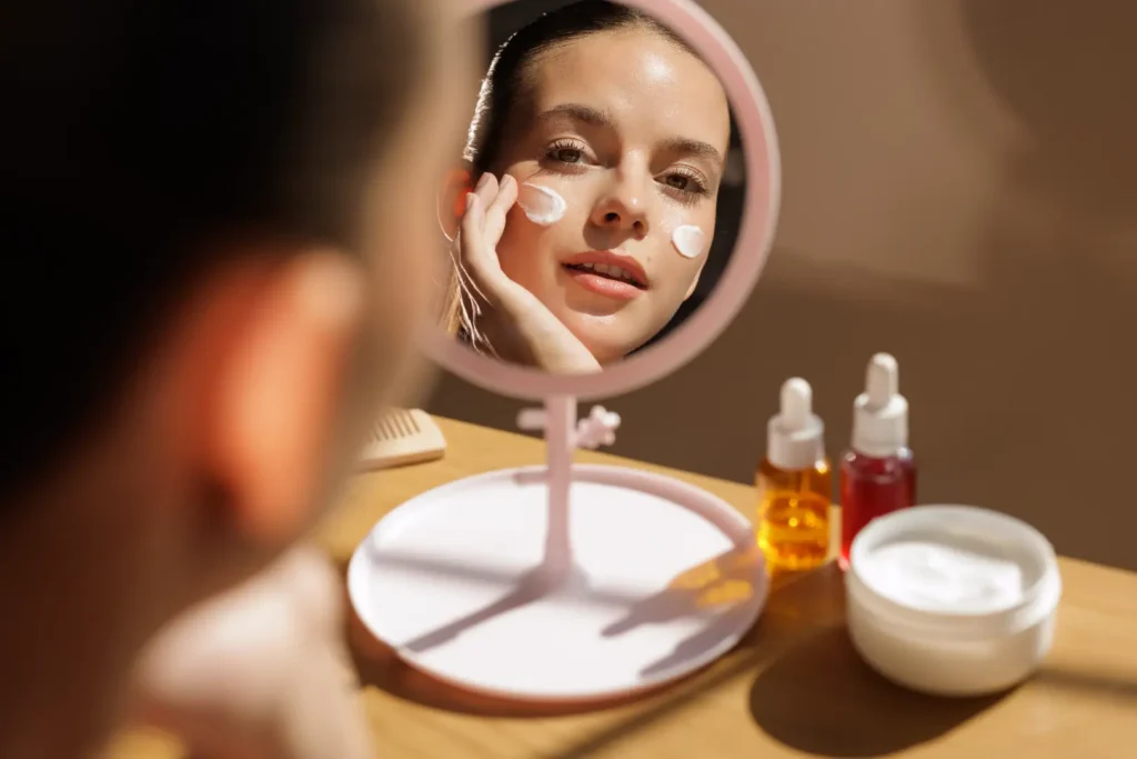 Woman looks into a tabletop mirror while applying white cream to both cheeks beside skincare products.